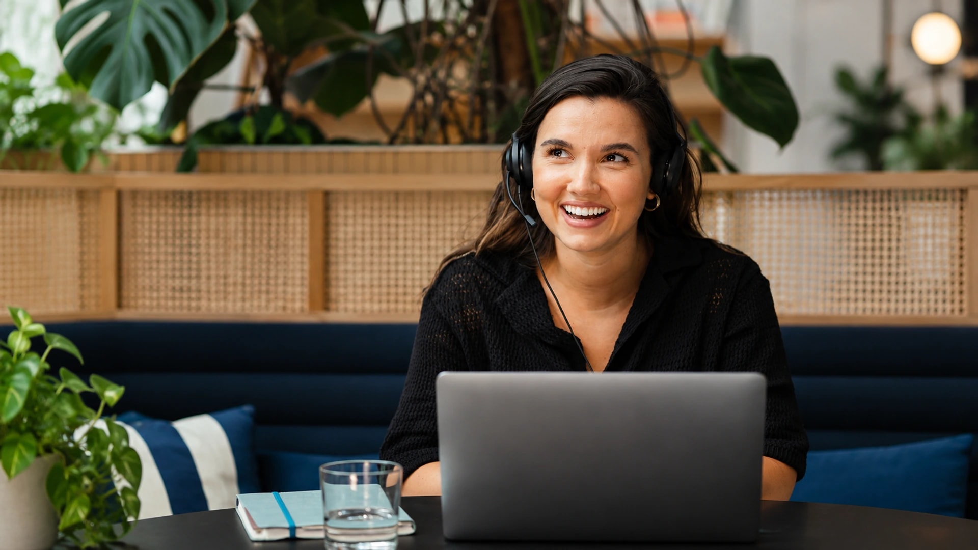 Mulher sorridente com headset diante de um notebook, em um ambiente com plantas.